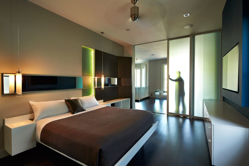 Primary bedroom features sliding, smoked glass entryway and striking use of light sources and color on wall details. Dark wood panelling, embedded colored lighting, hung cylindrical chandeliers, and a mirrored strip add texture and tone to wall above brown and white bed with matching white furniture.