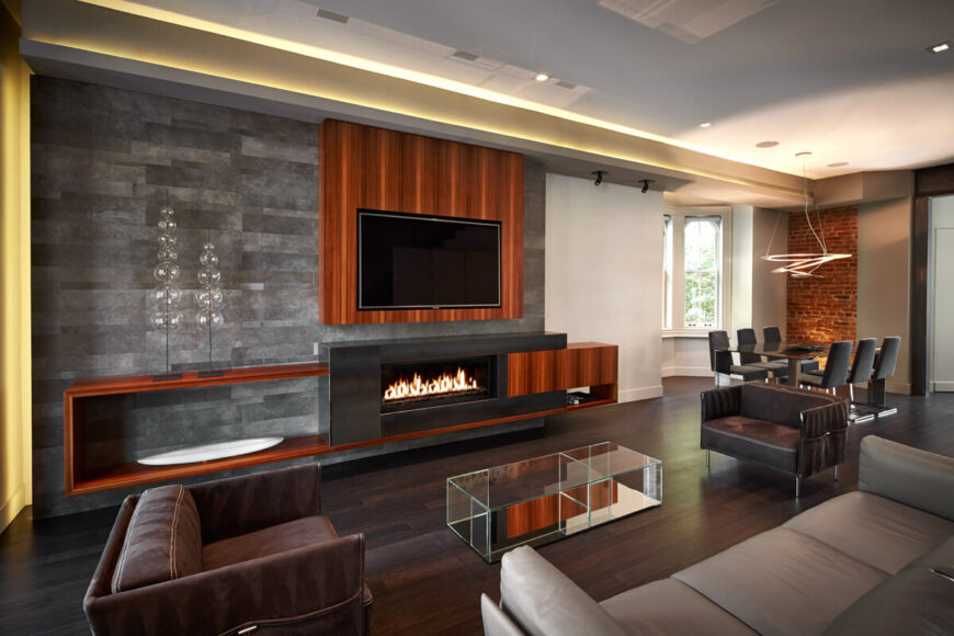 This is a more direct shot of the living room, showcasing the bright natural wood tones on wall supporting mounted television, with gas fireplace built into metal and wood shelving. Dining area is seen in background, with leather furniture up close.