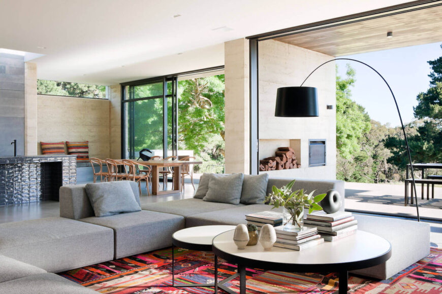 Central open living room space pictured here, with large grey U-shaped sectional centered around tiered circular coffee tables, over brightly colored area rug. Floor to ceiling glass affords view to outdoor space, while dining room and kitchen are pictured in background.