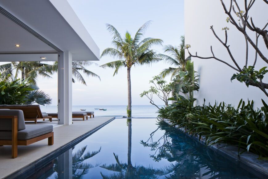 View from one of the bedrooms, overlooking the infinity pool toward the ocean. Rich wood and grey cushioned outdoor furniture sets feature on patio at left.