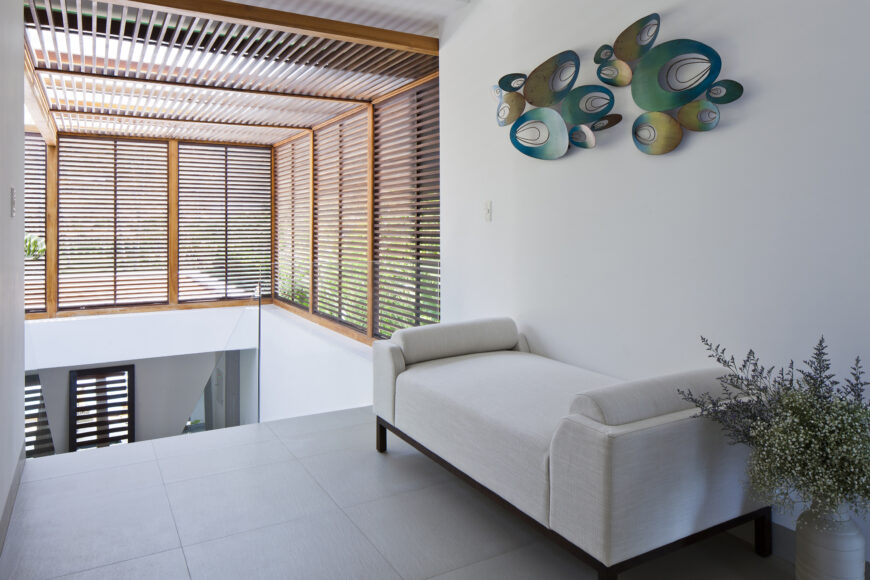 Upper level hallway features dark framed, white backless sofa integrating with the bright neutral tones. Sunlight filters through the louvers at left.