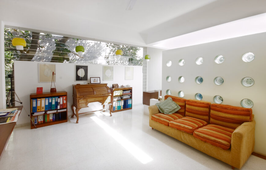 Upper level room featuring bright splashes of color, breaking up the natural wood on white theme. Orange sofa pairs with wood roll-top desk beneath multi-colored dome chandeliers, before expanse of upper wall exterior glass.