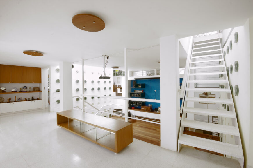 View overlooking the central living space, revealing separate staircases leading to each of the twin upper structures. Mixture of white tile and minimalist, natural wood textures defines the look.