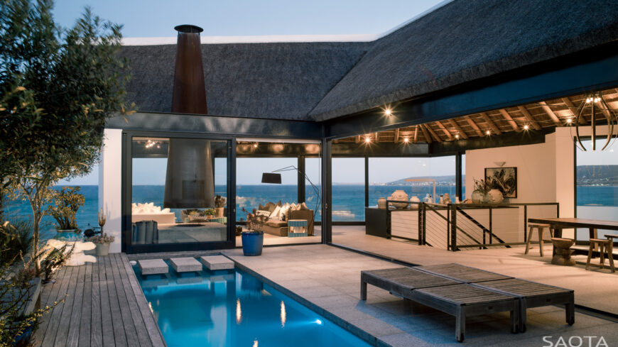 Viewing the central courtyard, we see a mixture of natural lighter tones splayed out beneath the natural wood beams and embedded lighting of the central structure. The pool separates the stone and wood surfaces, while full height glass reveals the interior throughout.