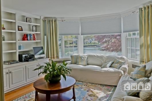 Another, smaller sitting room with a bay window and built-in shelving. Pastels in the rug and accent pillows add a soft pop of color to the white sofas and shelving.