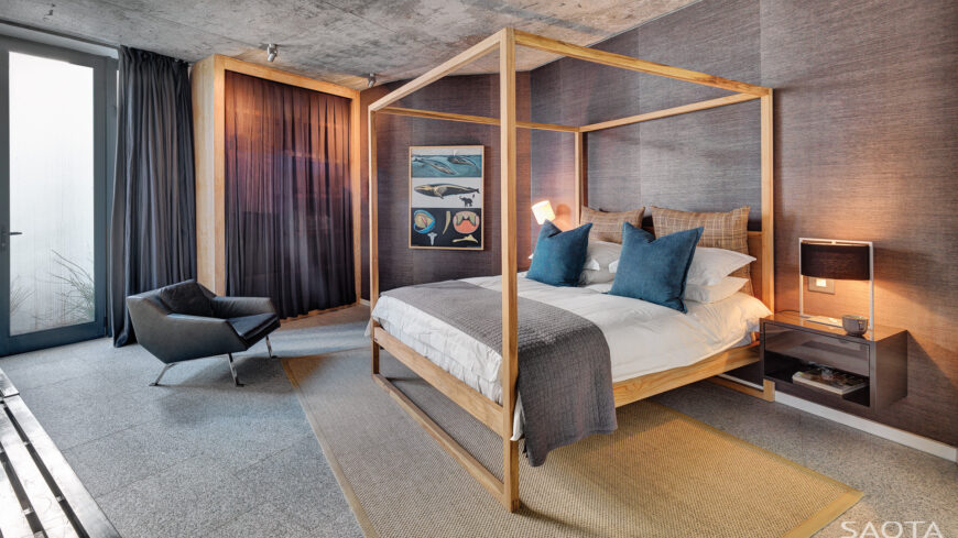 A second bedroom, pictured, holds floating bedside tables next to the natural wood frame bed, in a corner space with a solitary beige area rug over the marble flooring.