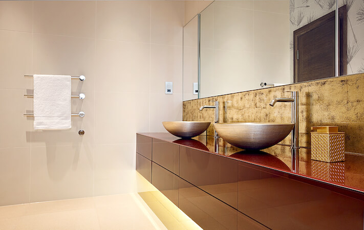 Another bathroom evokes an ultra-modern style akin to that of the kitchen, with sleek red cabinetry below a pair of stainless steel vessel sinks. Under-cabinet lighting highlights the soft white tone of the floor and wall tiles.