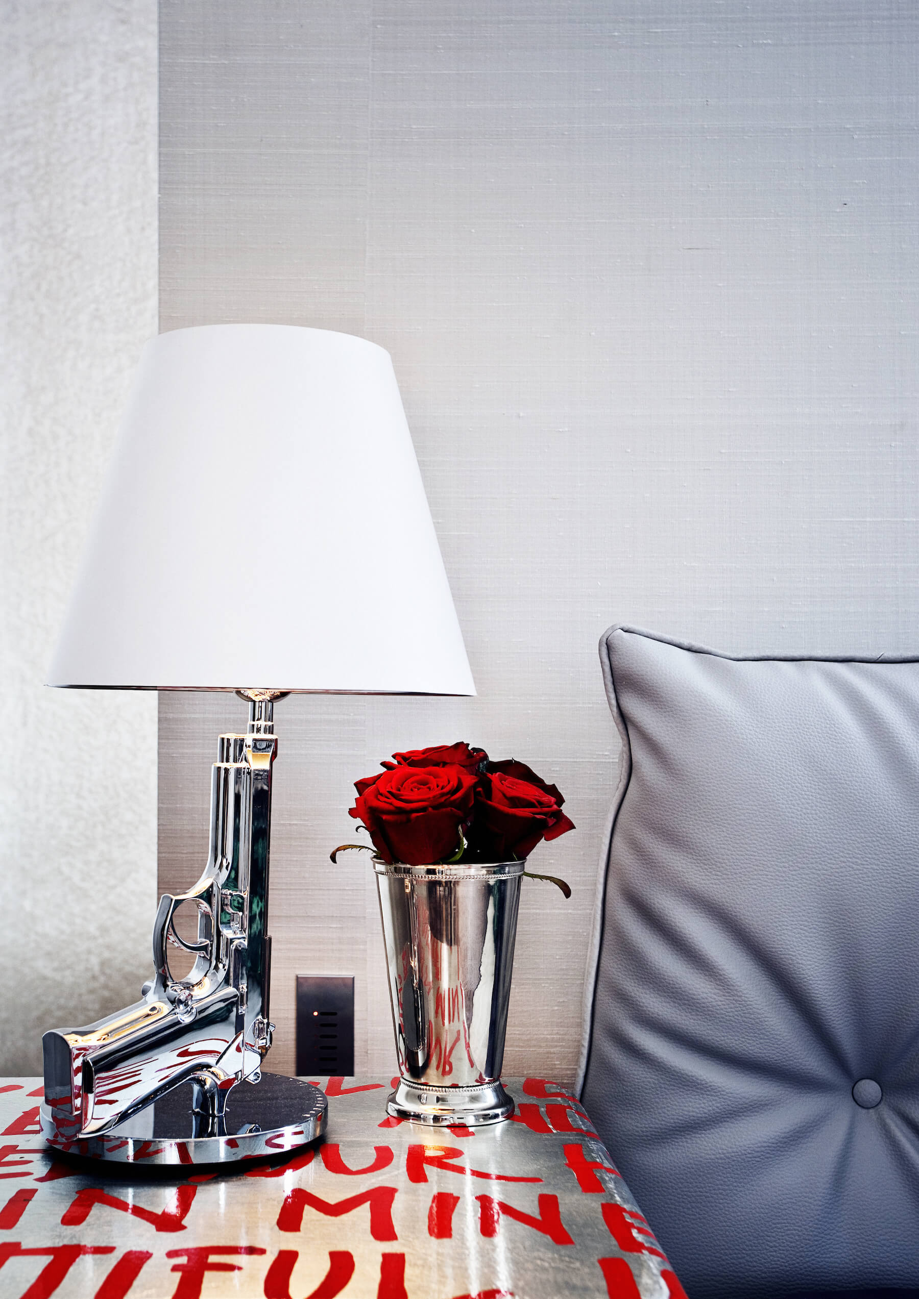 Silver toned bedside dresser with red text painting holds a chromed vase and this striking handgun-design lamp.