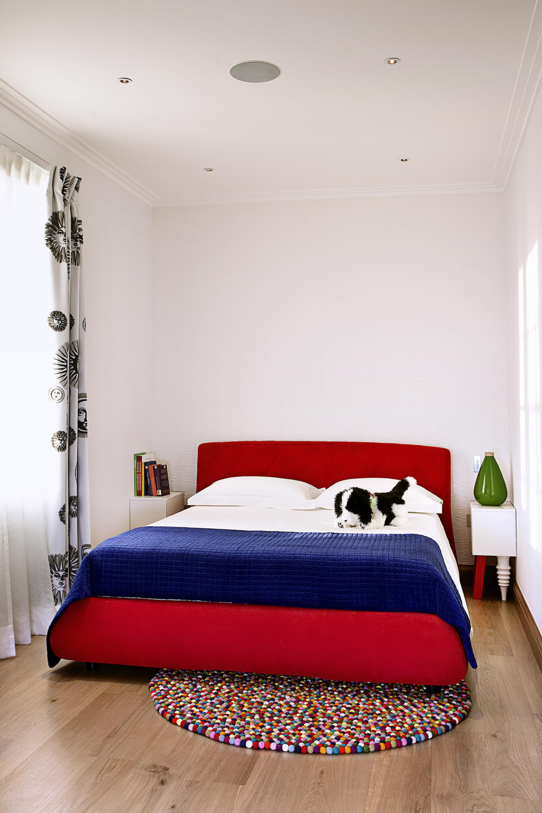 In this bedroom, we see another rainbow hued gum ball rug beneath a large red framed bed with blue and white sheets. The bed is flanked by a pair of white tables with mismatched legs.