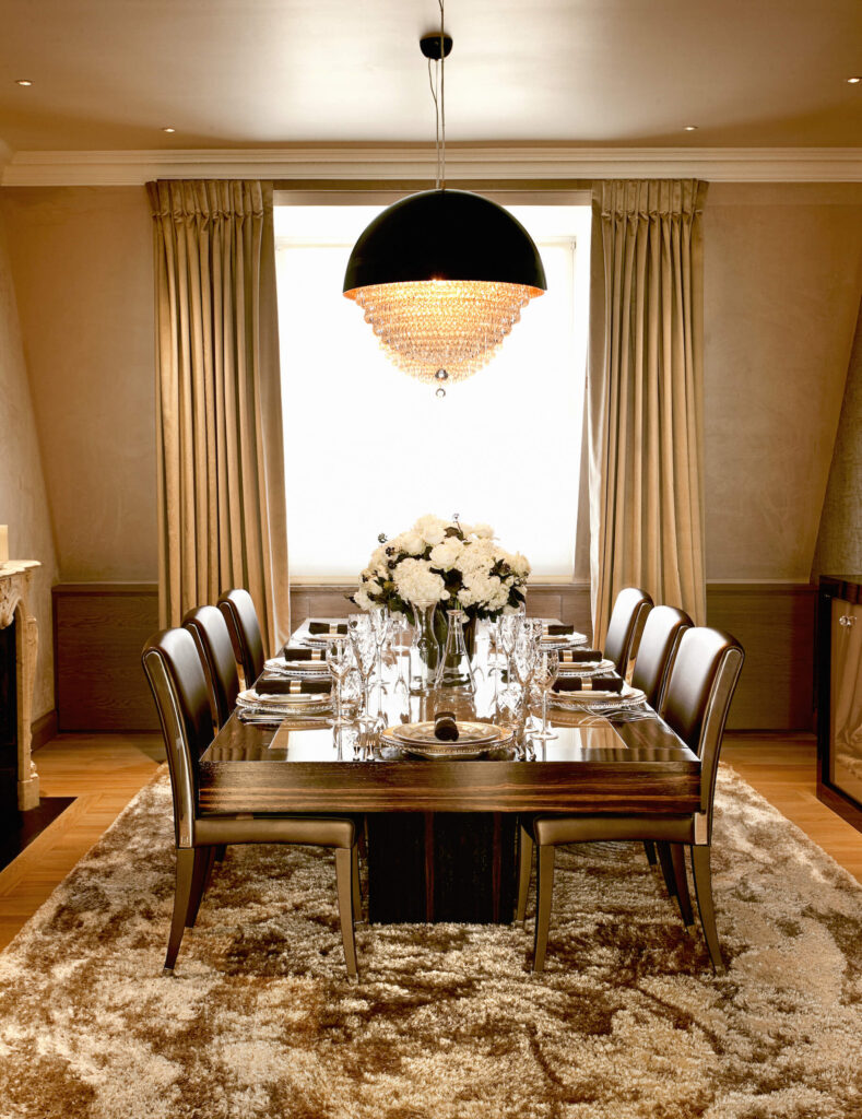 Dining room holds this extra thick wood dining table, paired with a set of leather upholstered side chairs beneath a unique chandelier combining a spherical body and traditional crystal structure. Gold hued drapes accent the rich tone of the room.