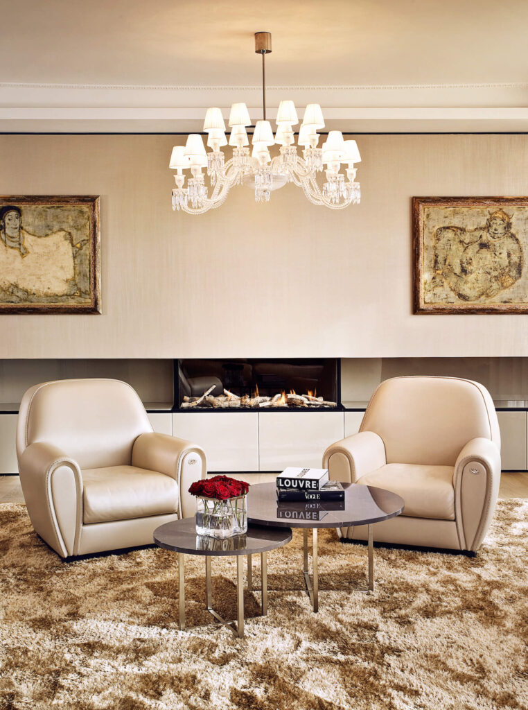  The bespoke living room combines traditional opulent details, like the chandelier and pair of cream leather club chairs, with a horizontal array of sleek minimalist cabinetry and open design fireplace. A pair of marble topped coffee tables stand over the thick brown rug on the hardwood flooring.