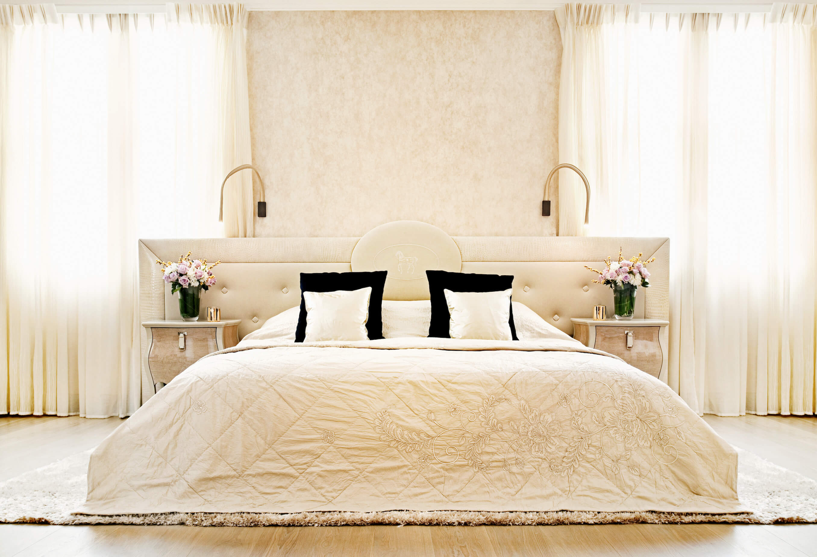 Primary bedroom suite is awash in bold cream tones, from the drapes to the large, button tufted headboard, flanked by a pair of carved wood dressers. Another thick area rug holds the furniture in this room over hardwood flooring.