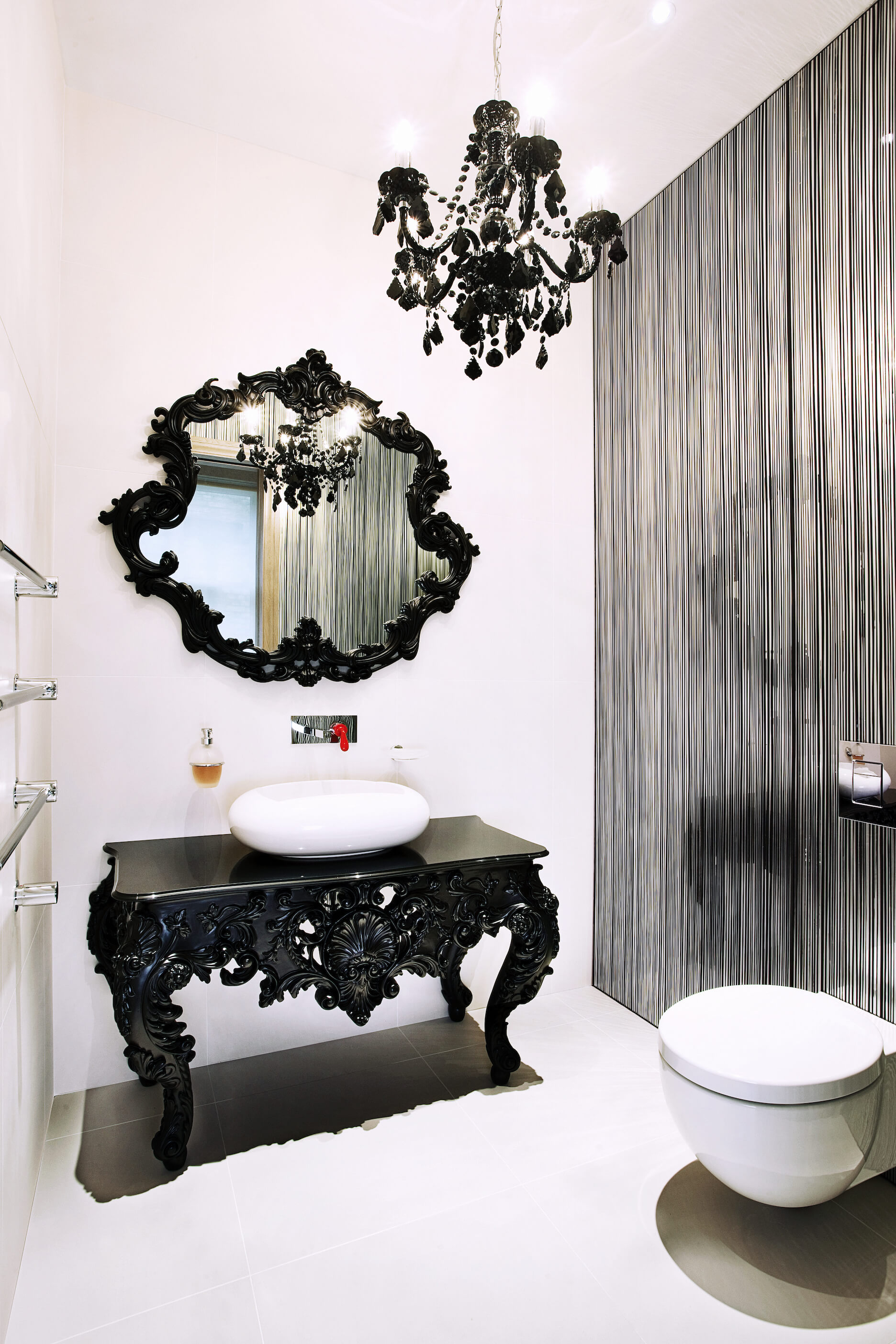 Finally, we have another completely distinct bathroom. This example sports an ornate, traditionally styled table vanity in black with white vessel sink, below a complementary black framed mirror. Silver vertical stripe wall contrasts with white tiling throughout.