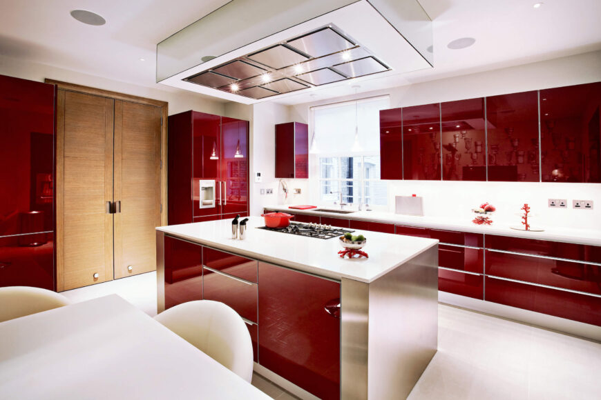 The red modern kitchen leaps out with grand contemporary style, holding red glossy cabinetry and stainless steel island with white flooring and countertops. This look contrasts deeply with the living room, offering a fresh sense of luxury in the space.