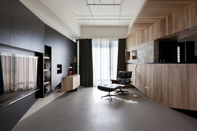 Large format tile flooring spread out beneath the entire open space. Minimalist dark wood cabinetry at left provides an angled, sleek structure in contrast with the lighter wood of the kitchen, and sunlight via the full height window at back.