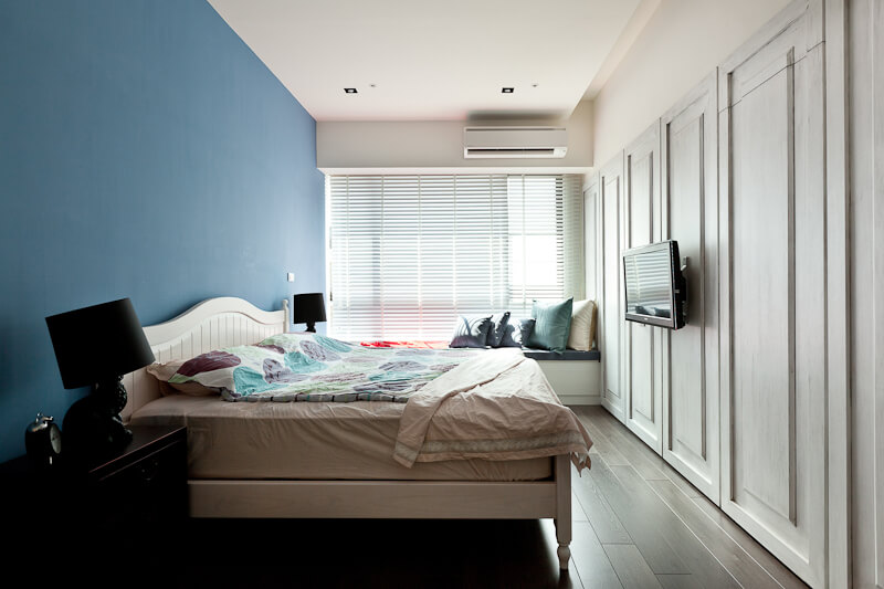 The primary bedroom features dark hardwood flooring, wall-length panels in white, and a bold blue wall behind the wood frame bed. Full height window grants natural light throughout. 