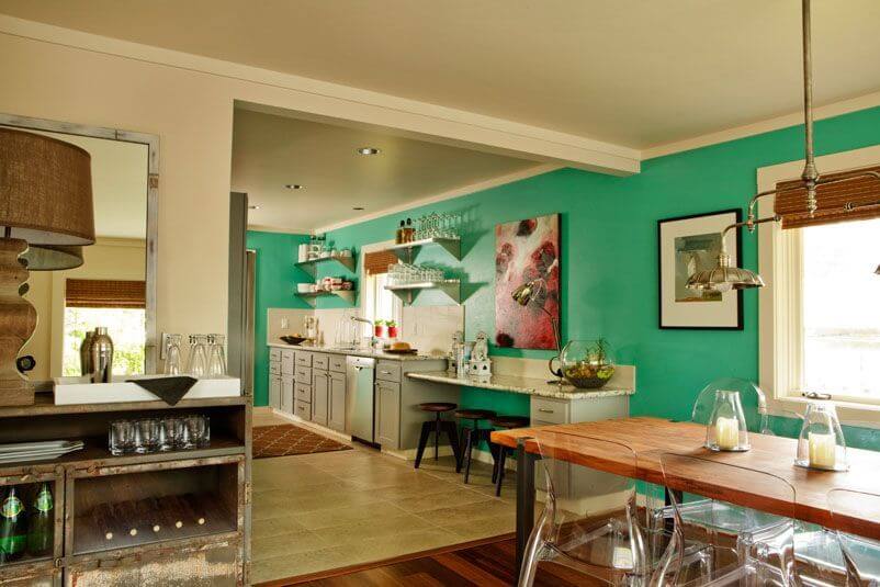 The open-concept dining room and kitchen are in a bold blue-green. The dining set is wood with transparent chairs. The kitchen is in a light beige tile with a white backsplash.