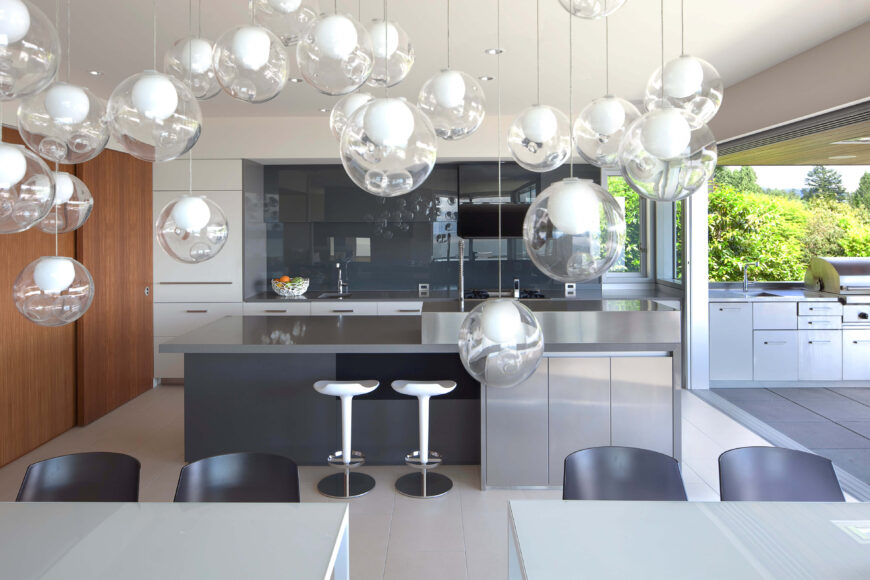 Across the divided dining table, we see the kitchen, highlighted by a large island in sleek brushed metal. A cloud of glass pendant lights hangs in the foreground, reflected in the glossy backsplash.