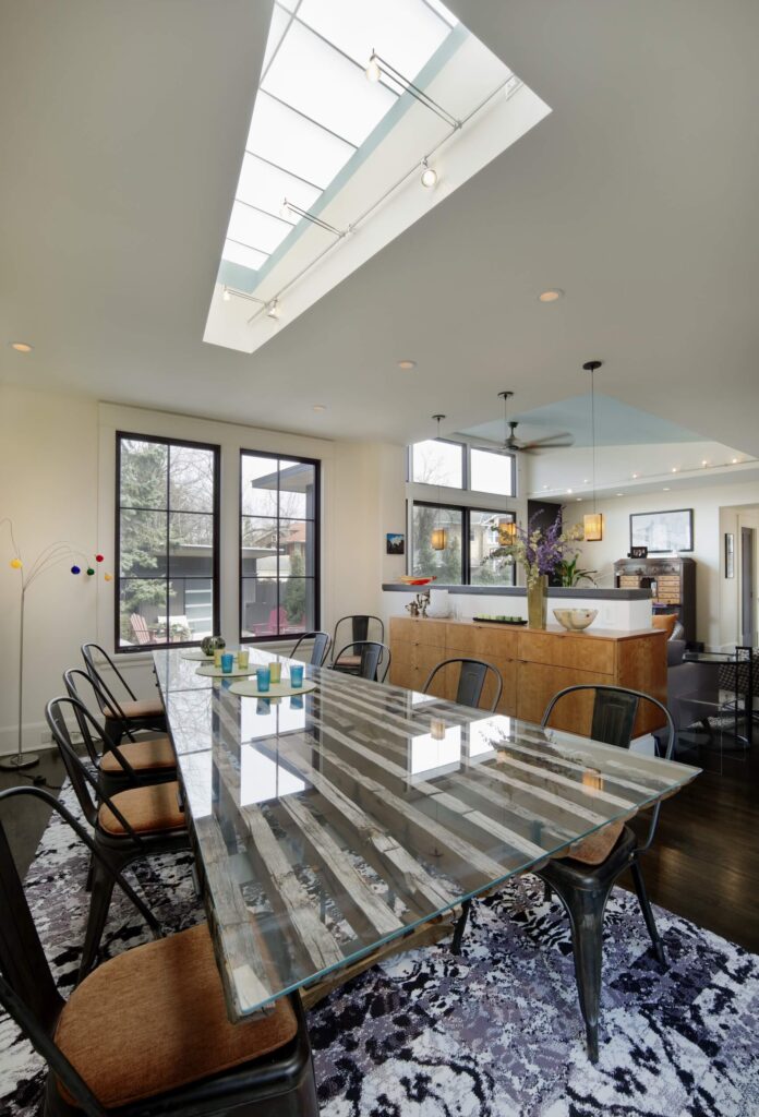 Dining area features a glass table framed with natural wood and paired by cushioned chairs over a shaggy rug. It also has a skylight with industrial lights.