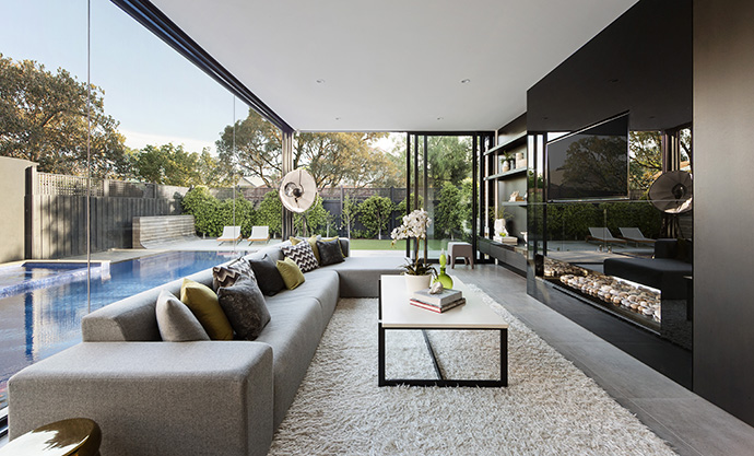 The living room sits beside the pool outside, and a dark glossy entertainment center reflects onto the landscaping. A contemporary color scheme in the furniture matches the color of the flooring and walls of the home.