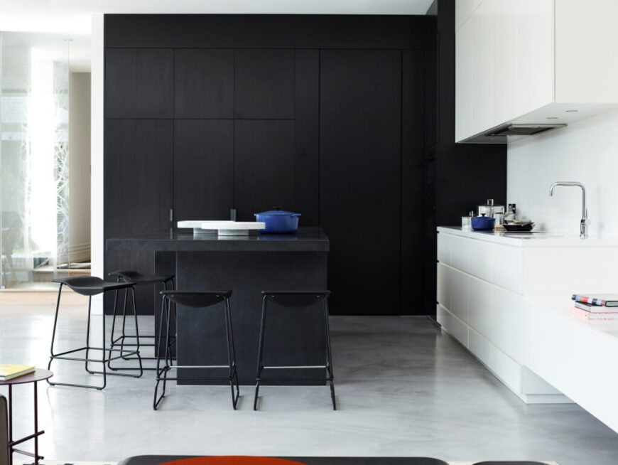 From here we see the high contrast defining this corner of the open-plan space. Sleek white cabinetry surrounds the sink and matching countertops, while an imposing set of floor to ceiling dark wood cabinets fills the remaining wall space.