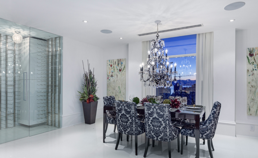 The dining room takes advantage of dark accents and bold damask patterns to make it stand out from the rest of the decor. To the left of the room is the gorgeous climate controlled glass wine storage. The stunning crystal chandelier over the table continues the use of dark accents while balancing out the brightness of the room.