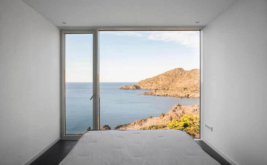 We enter one of the home's minimalist bedrooms, which also features a glass-paneled door that leads outside. Storage is kept behind the bed so that it doesn't obstruct the view of the craggy coast.