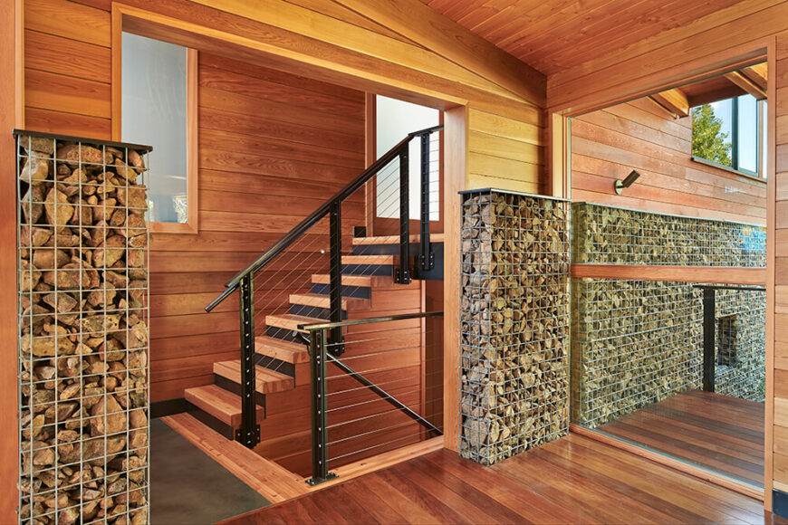 Details such as continuing the use of gabion walls indoors and utilizing sturdy, industrial railings keeps with the rustic, camp feel achieved by the house. This shows off the gorgeous wood paneling and flooring used in the living areas.