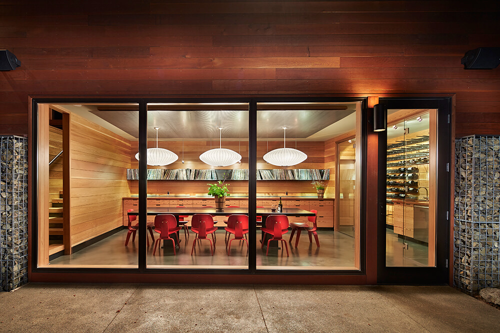 Warm dining room surrounded by shiplap walls and accented by vibrant red chairs. It has a lengthy dining table lighted by large pendants. There's also a wine cellar enclosed with glass walls next to it.