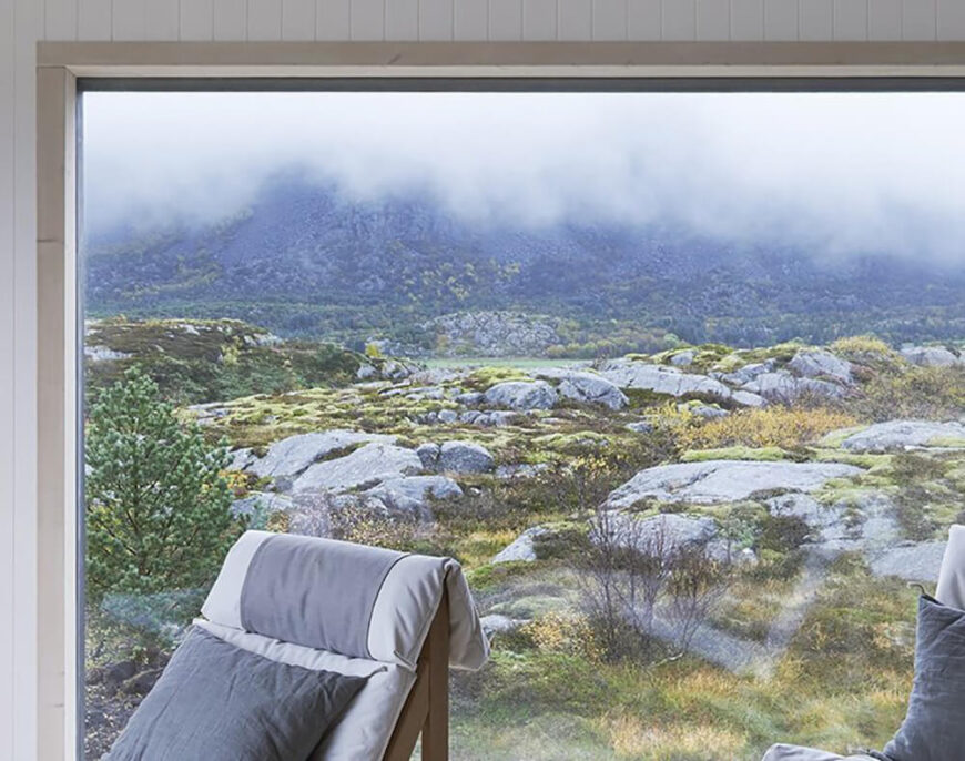 Over the natural wood chairs and through the vast living room window, se can see the mountains rising in the distance, across acres of rocky terrain. The windows are framed in light natural wood for high contrast.