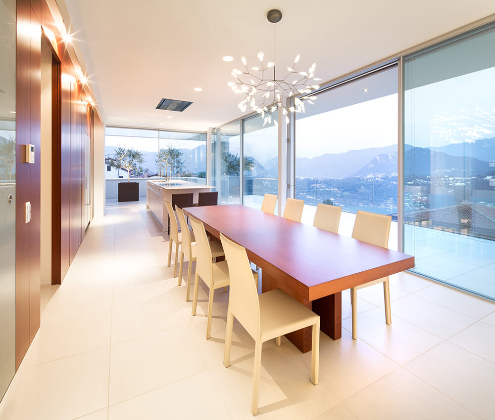 Enjoy a mesmerizing mountain view through the glass walls of this dining area. It has a long wooden table with white chairs and lighted by a fancy chandelier.