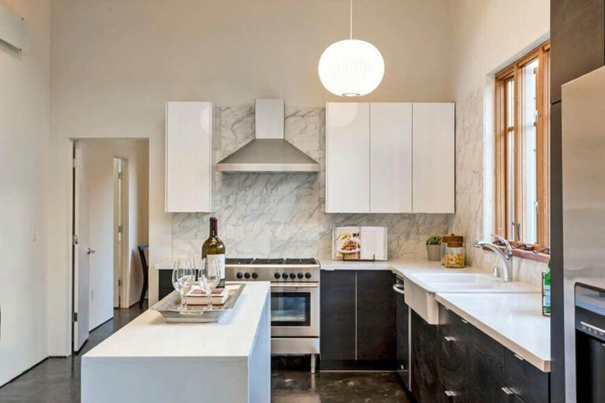 Here's a close look at the kitchen, with white countertops, black cupboards, and a bold marble backsplash that wraps the upper white cabinetry and sink area. A slim white island offers more contrast and helps define the area.