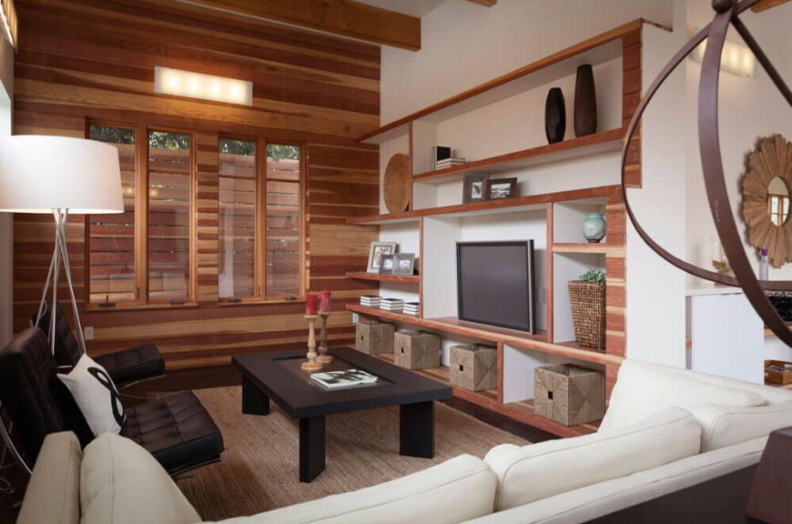 At the far end of the open area, we see the living room decked out in swaths of natural wood and elegant contemporary furniture. A complex set of built-in shelving on the wall provides space for art and storage, while the all-wood feature wall at left continues this element within the home.