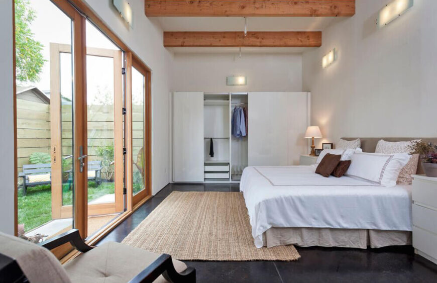 The primary bedroom is awash in white, contrasting with the dark flooring that appears throughout the home. In here we see more of the exposed wood beams, as well as a large set of wood framed glass French doors with direct access to the backyard.