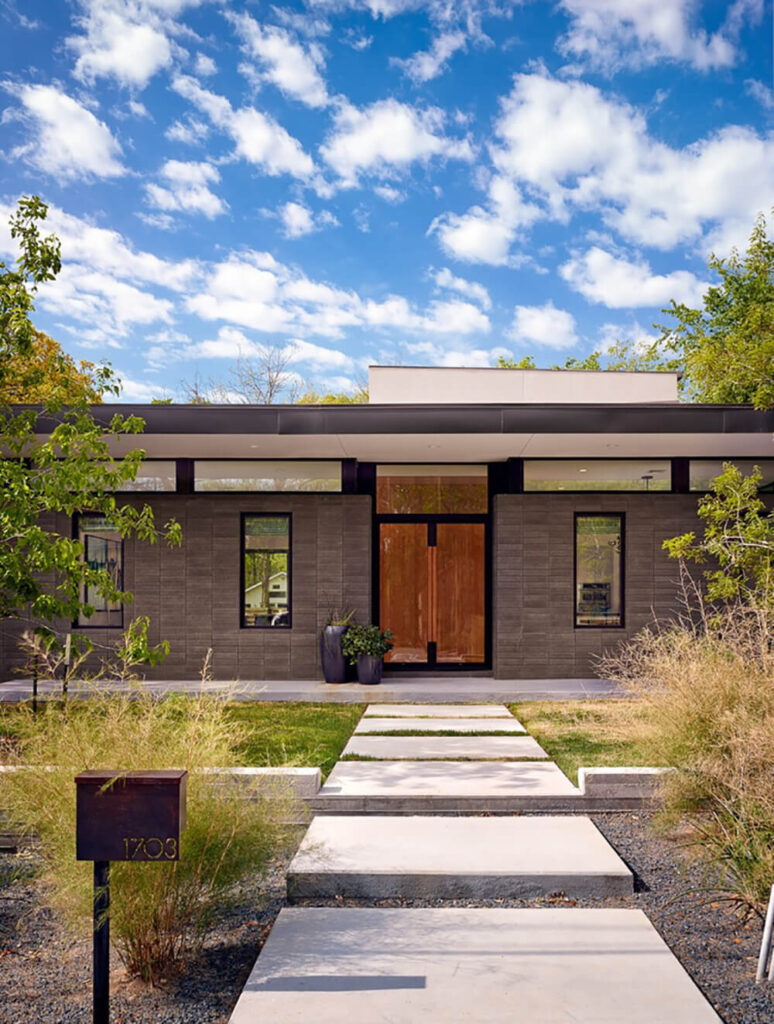 Across the large stone slab walkway we see the unassuming home, with slimmer windows on the front and a low, broad flat roof obscuring the brightly open space within.