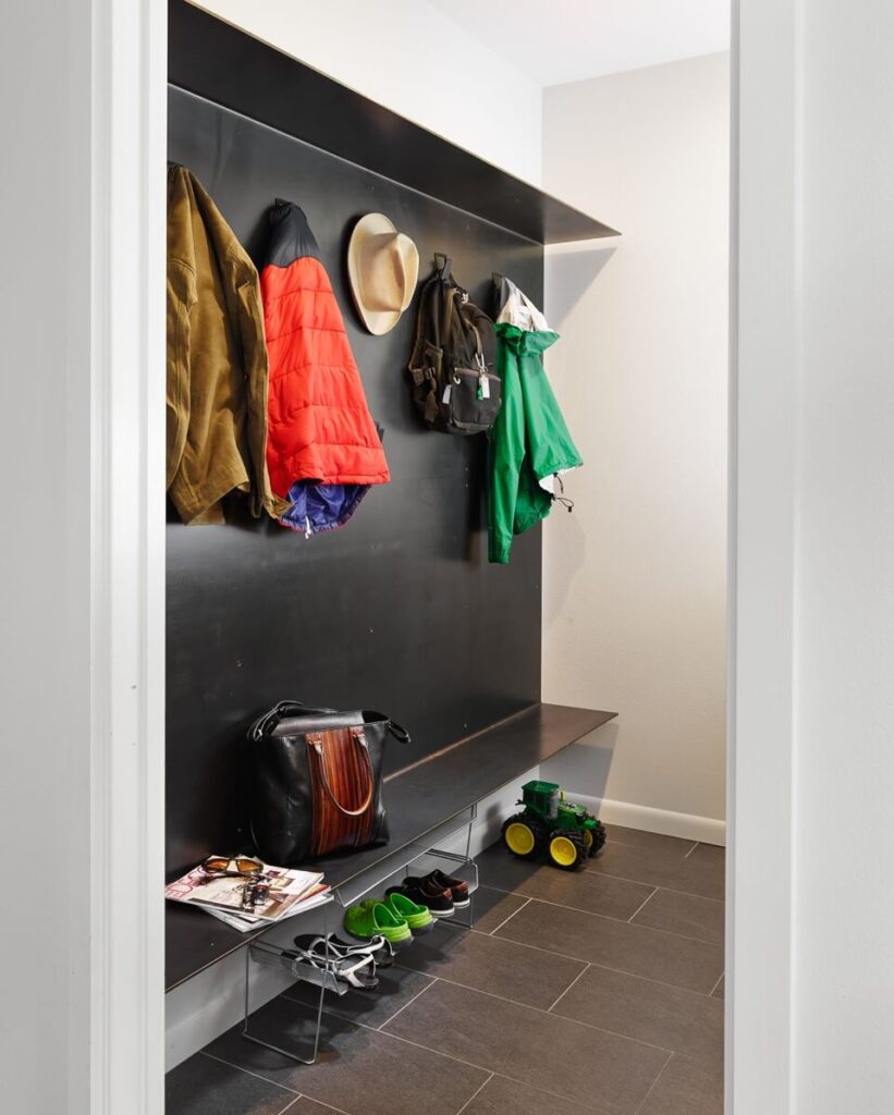 The mudroom features dark tile flooring and a unique metal wall feature, with a built-in shelf. This simple, open place for shoe and coat storage exemplifies the ethos behind the home design.