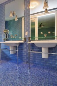 Blue-on-blue primary bathroom features mosaic tile floors and brick-pattern tile walls with a freestanding tub and two floating sinks. Wall-mounted lights are installed in large mirrors over the sinks.