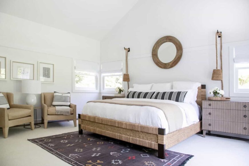 Clean, white primary bedroom designed with round wooden wall mirror placed above the bed and a printed black rug. It has beige armchairs with a table lamp in the middle. 