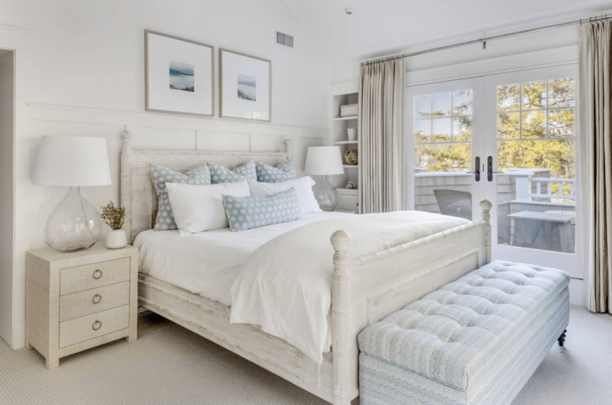 Charming primary bedroom with wooden bed and a lovely blue tufted bench on its end. It includes wooden nightstands with round glass table lamps and carpet flooring.