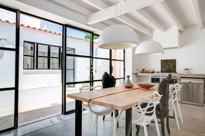 The glass walls of this white dining room give it an illusion of being outside the house. The natural lighting brightens up the white ceiling with exposed wooden beams and white teardrop dome lighting. The wood-top dining table with a division in the middle and the surrounding assortment of chairs give it an informal air and warmth.