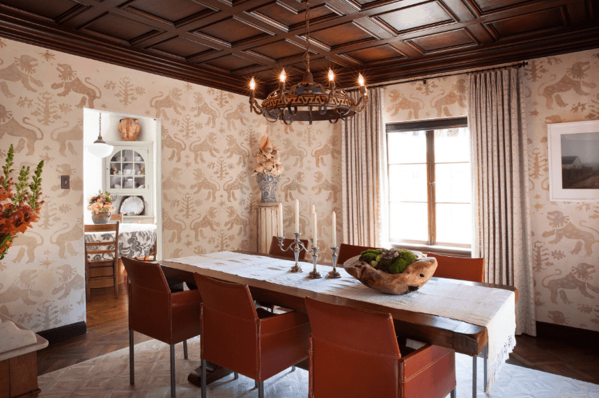 The elegant dark wood finish of the ceiling and the matching dark brown chandelier accentuates the patterned walls and dark wooden floor. The long slab table is surrounded by leather vintage chairs and topped with a wooden centerpiece and a table runner that matches the area rug.