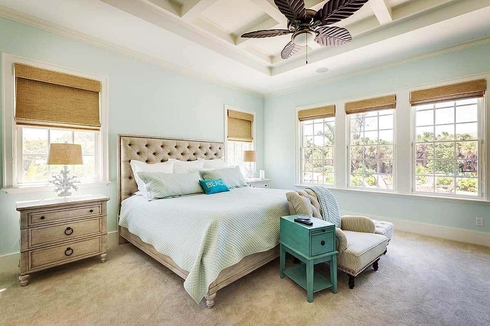 A comfy tufted bed, light wood nightstands and a striped armchair flanked by a green side table and ottoman fill the primary bedroom. These are then complemented by the coffered ceiling and glass windows that bring in natural lights.