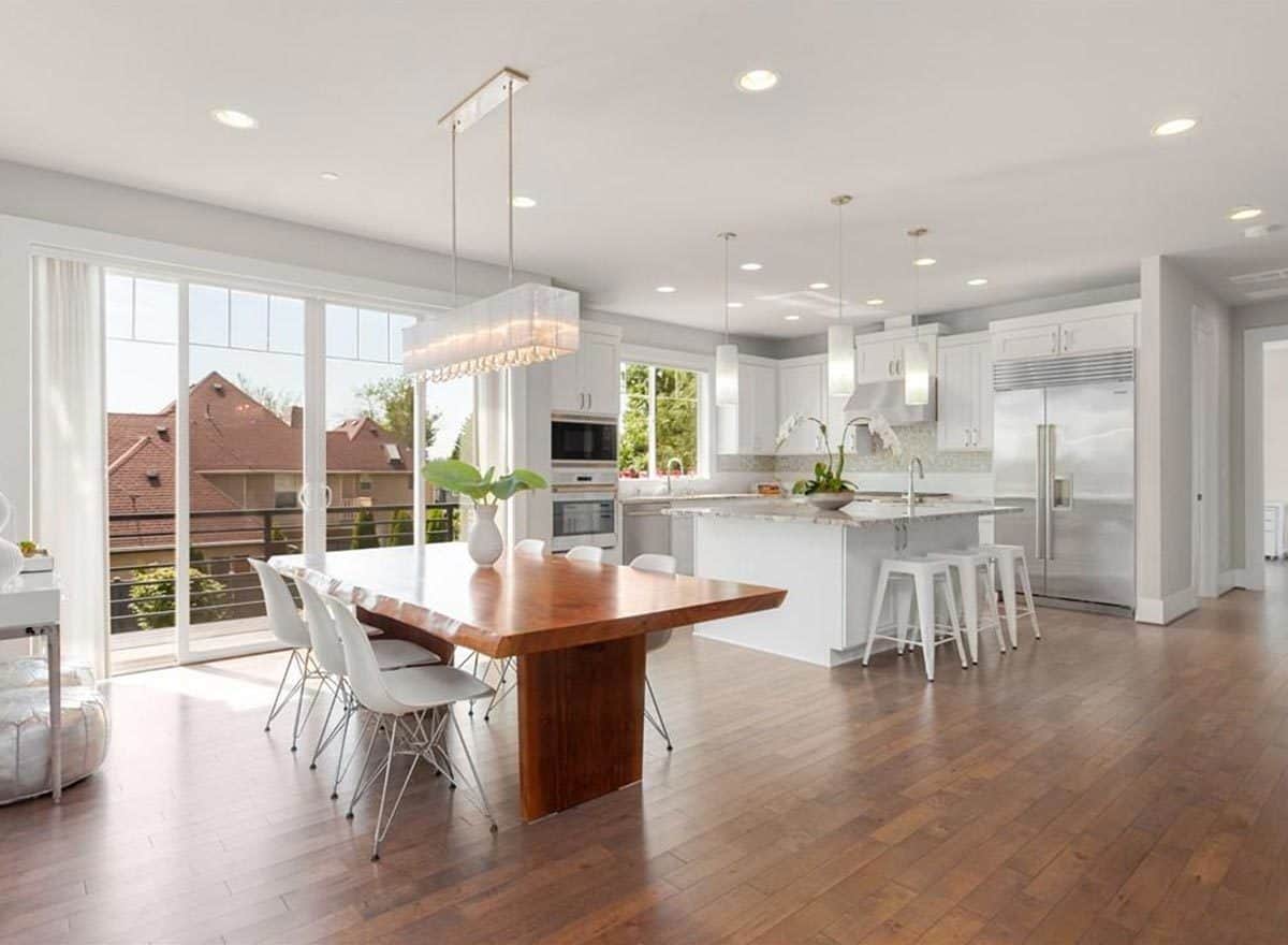 An open dining area with white modern chairs and a large wooden dining table topped with a linear chandelier. It flows right into the kitchen brightened by recessed ceiling lights and cylindrical pendants.