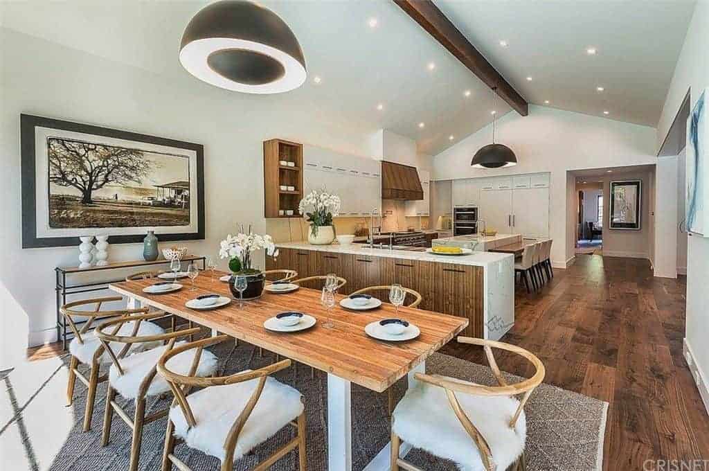 The beautiful painting of a farm with a large tree gives a quality of warmth and home to the white wall by the head of the wooden dining table with matching armchairs that has furry seat cushions. This is topped by a large black dome pendant light hanging from the single exposed beam of the cathedral ceiling.