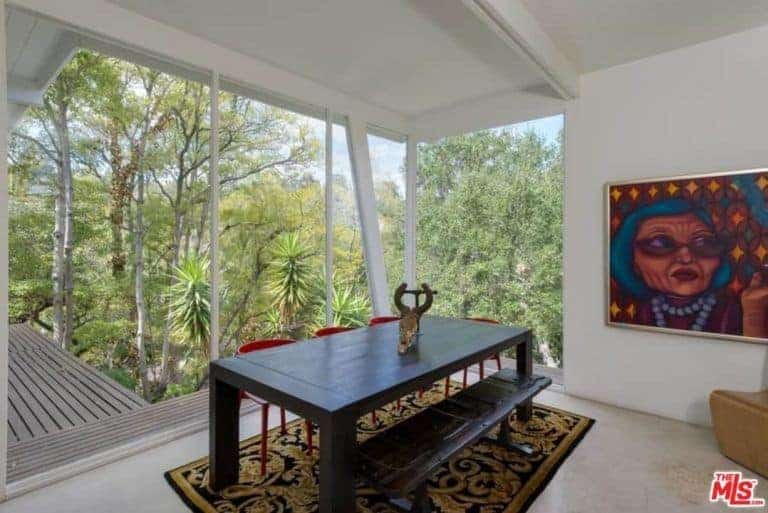 The highlight of this medium-sized dining room is the lush landscape of tall trees featured by the glass walls that are framed with white ceiling, walls and flooring that contrasts the dark wooden dining table and bench over a colorful patterned area rug.