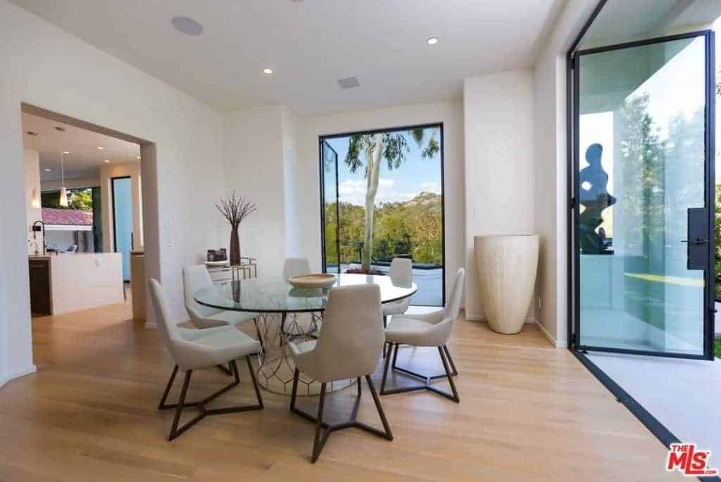 The two adjacent walls that open up due to the tall glass doors let in an abundance of natural lighting that makes the glass-top of the modern round table shine and makes the light hardwood flooring brighter as well as the white walls and white ceiling complemented by the light gray cushioned modern chairs.