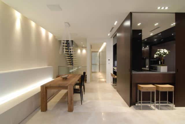 This bright medium-sized dining area beside the kitchen has beige marble flooring and beige walls that has a built-in white bench paired with the wooden dining table. There are modern accents due to the back lighting of the walls and bench together with the recessed lights of the white ceiling.