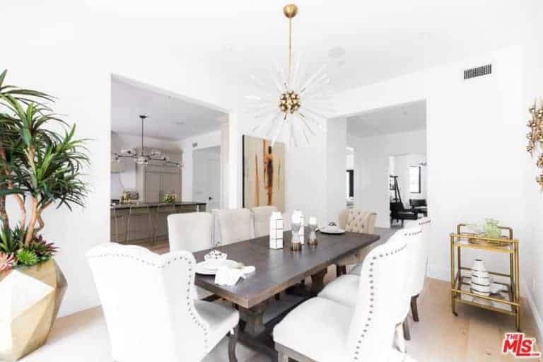 This medium-sized dining room has white walls, white ceiling and light hardwood flooring for that bright aesthetic that is augmented by the white cushioned dining chairs that stand out against the dark wooden rectangular dining table. This also makes the golden elements stand out like the pendant light, tray trolley and the pot of the plant.