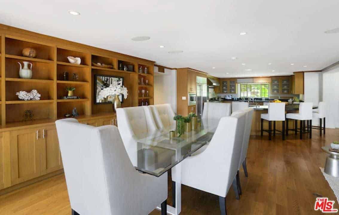 An entire wall of this dining area by the kitchen is dominated by a large wooden structure that has built-in cabinets and shelves. This structure matches the tone of the hardwood flooring and contrasts the white ceiling that has recessed lights over the glass-top rectangular dining table and its light gray cushioned dining chairs.