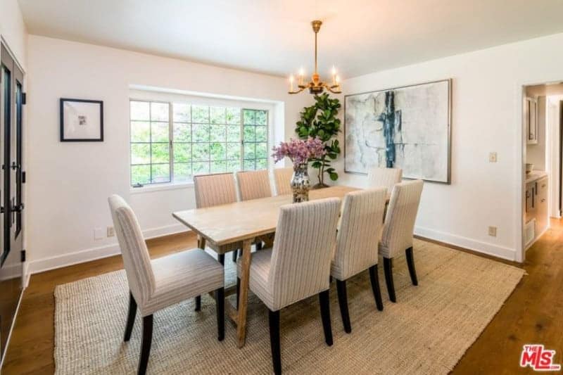 The rustic woven area rug that complements the hardwood flooring matches well with the Farmhouse-style wooden dining table. This is surrounded by dining chairs that have striped gray cushions that complement the bright walls and ceiling that hangs a simple chandelier over the table.